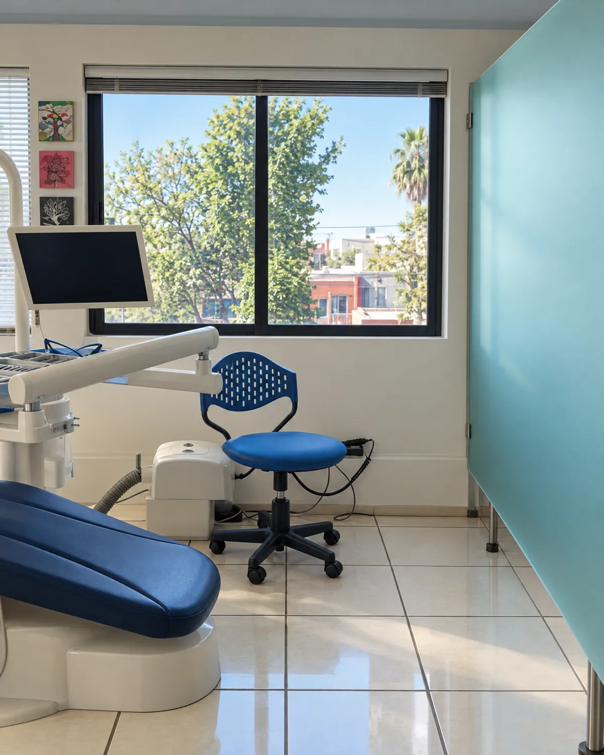 Treatment room: blue dental chair, stool and a window with trees, next to a turquoise frosted-glass partition