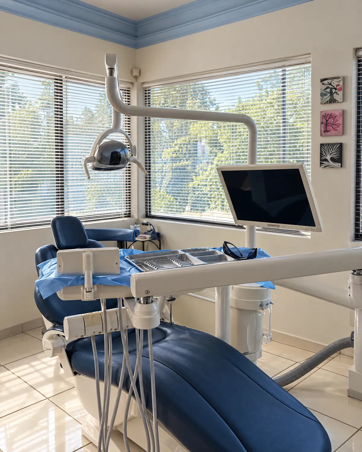 Blue dental chair with lamp and monitor, between two windows with blinds